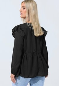 Blonde nainen pukeutuneena löysään mustaan paitapuseroon, jossa on frilloja hartioilla, ja vaaleansinisiin farkkuihin, seisoo sivuliikkeessä tasaisella taustalla.