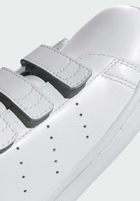Baskets en cuir blanc avec deux bandes Velcro, bout rond, petites perforations sur le côté et semelle en caoutchouc texturé.