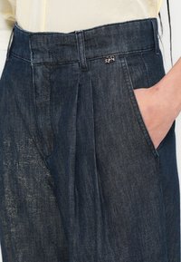 Sötét farmernadrág, magas derékkal, redőkkel és oldalzsebbel, finom fényű, kis mintás díszítéssel a derékrészen.