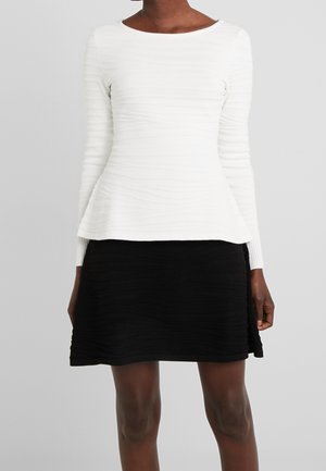 Femme portant un haut texturé blanc à manches longues et une jupe texturée noire longueur genou sur fond gris.
