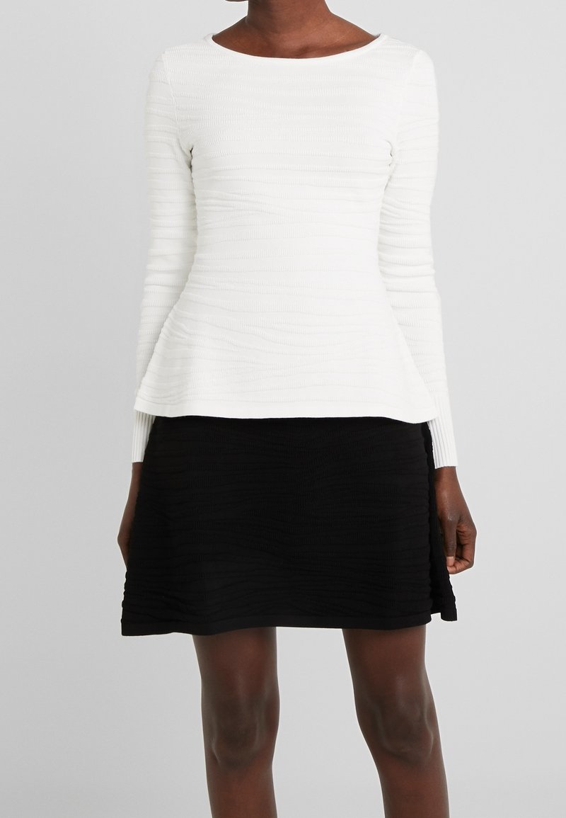 Femme portant un haut texturé blanc à manches longues et une jupe texturée noire longueur genou sur fond gris.