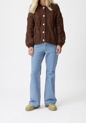 Mujer vistiendo un cardigan de punto grueso marrón con botones de perla, jeans rectos de color azul claro y zapatos gruesos beige, de pie.