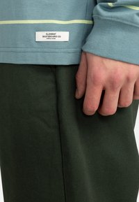 Langärmliges Hemd aus Baumwolle in Hellblau mit gelben Streifen, kombiniert mit grünen Hosen. Das Etikett auf dem Hemd lautet "ELEMENT SKATEBOARD CO."