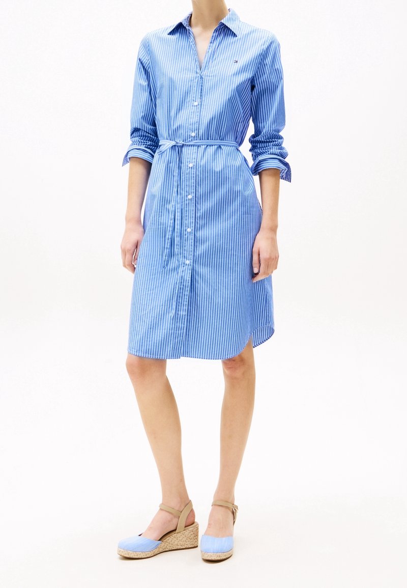 Femme portant une robe-chemise à rayures verticales bleues et blanches avec manches retroussées et sandales compensées bleu clair.