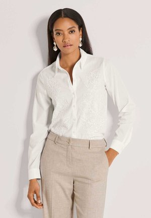 Frau trägt eine weiße Bluse mit Knöpfen und Blumenstickerei sowie beige, hochgeschnittene Hosen, steht mit einer Hand in der Tasche.