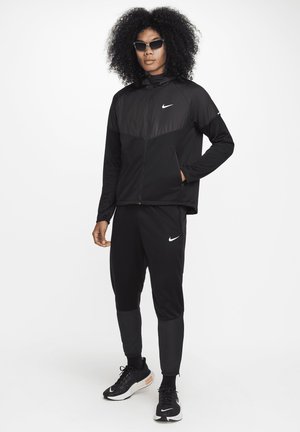 Black athletic jacket with a zip front and long sleeves, paired with matching joggers. Features Nike logo in white. Worn with black sneakers.