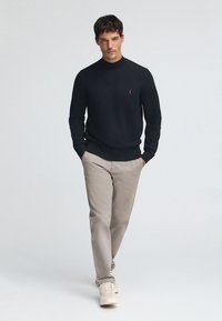 Svart stickad tröja med rund halsringning, kombinerad med ljusgrå chinos och cremefärgade sneakers. Märkbart rött logo på tröjans bröst.