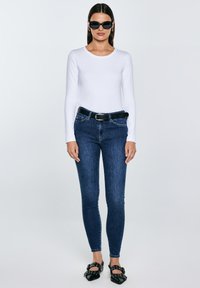 Žena koja nosi crne sunčane naočale, bijelu bluzu s dugim rukavima, tamnoplave uske traperice s crnim remenom i crne špicaste cipele sa remenčićima.