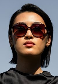 Bruine oversized zonnebril met een schildpadmotief frame, donkere gradient lenzen en geometrische uitsparingen; gedragen met een zwarte satijnen top.