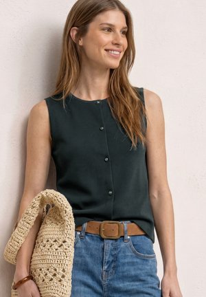 Kvinde smilende og kigger mod højre, iført en sort ærmeløs knapbluse, blå jeans med brunt bælte, holder en beige strikket taske.