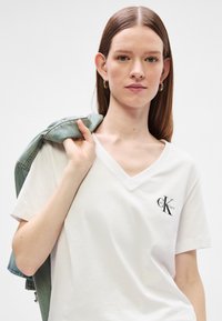 Biela bavlnená tričko s výstrihom do V, s čiernym logom CK na ľavej strane. Modelka drží džínsovú bundu cez plece.