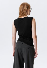 Top nero senza maniche abbinato a pantaloni grigi su misura; il tessuto è liscio con una vestibilità casual, caratterizzato da una leggera arricciatura sul retro in vita.