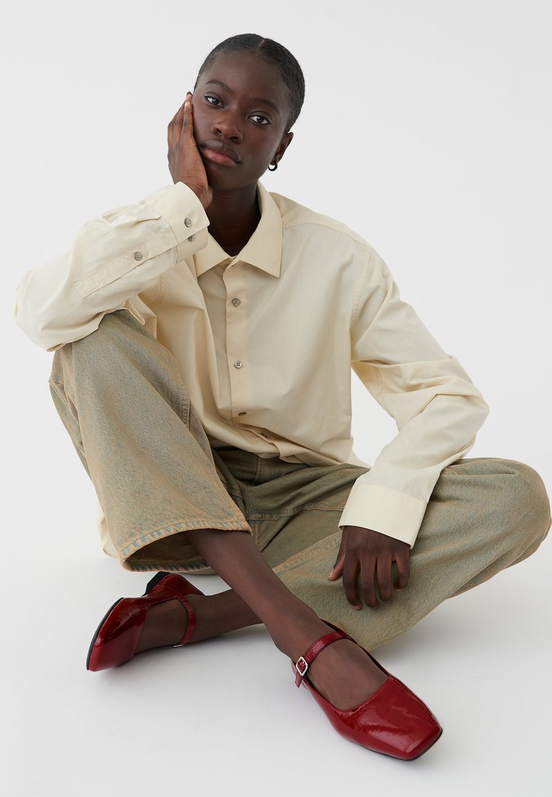 Beige knäppt skjorta, lösa gröna texturerade byxor, röda lackskinnskor med kvadratig tå och ankelsnodd. Modell sitter med handen på ansiktet.