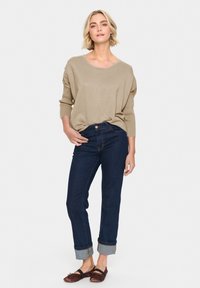 Maglione a maglia beige, vestibilità ampia, scollatura a barca, abbinato a jeans di denim blu scuro arrotolati alle caviglie. Ballerine marroni, texture liscia.