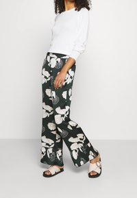 Biała długa bluzka z okrągłym dekoltem zestawiona z zielonymi szerokimi spodniami w kwiaty i beżowymi klapkami z okrągłą klamrą.