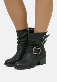 Bottes en cuir noires mi-mollet avec Talons blocs, fermeture éclair sur le côté, et trois boucles en argent sur des jambes lisses et brunes, le tout sur fond blanc.