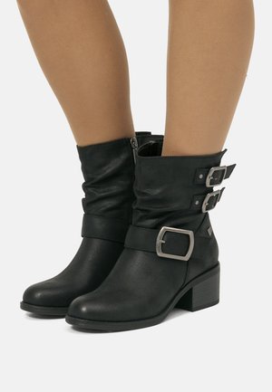 Bottes en cuir noires mi-mollet avec Talons blocs, fermeture éclair sur le côté, et trois boucles en argent sur des jambes lisses et brunes, le tout sur fond blanc.