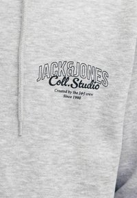 Sudadera gris de tejido suave con un logo bordado en negro, con textura elevada y detalle de cordón en la capucha.