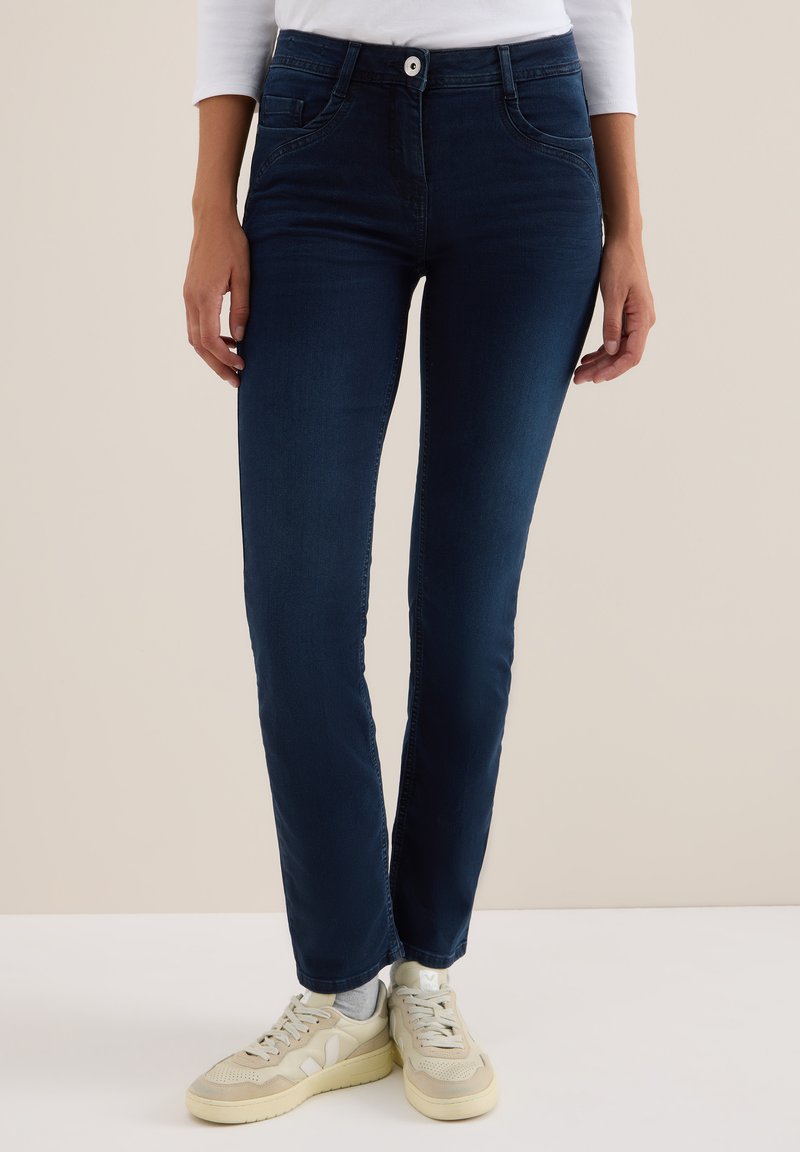 Jeans de mezclilla azul oscuro con un corte ajustado, que cuentan con bolsillos delanteros, cremallera y cierre de botón. Combinados con zapatillas de color claro.