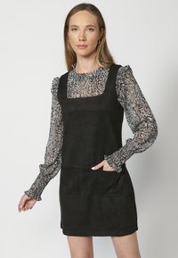 Robe en suede noire avec un décolleté carré, dotée de deux poches avant. En dessous, un chemisier transparent avec un motif floral noir et blanc.