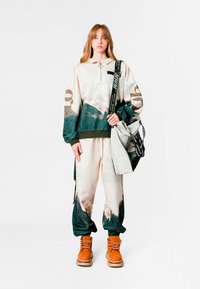 Sudadera con capucha que presenta un gráfico de montaña en beige y verde oscuro; combinada con pantalones a juego y botas naranjas; incluye un bolso tote texturizado.