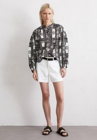 Blouse à motifs noir et blanc avec de longues manches bouffantes, un col haut, associée à un short blanc et des sandales noires avec des accents dorés.