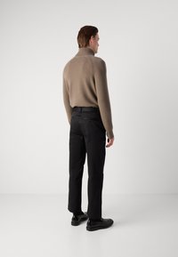 Beige strikket turtleneck sweater, ribbede manchetter, parret med sorte bukser og sorte sko, set fra bagfra mod en neutral baggrund.