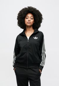 Must zip-up jope valgete kolme triibuga varrukaulidel ja väike valge Adidas logo rinnal. Kerge materjal ja vaba lõige.