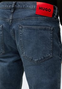 Μπλε τζιν από τζιν ύφασμα με ορατές πίσω τσέπες, με κόκκινη δερμάτινη ετικέτα με "HUGO" σε μαύρα γράμματα στη μέση.