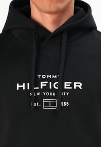 Sudadera negra con el texto y logo en blanco "Tommy Hilfiger New York City est. 1985" en el pecho, llevada por una persona desde el cuello hasta la parte superior del torso.