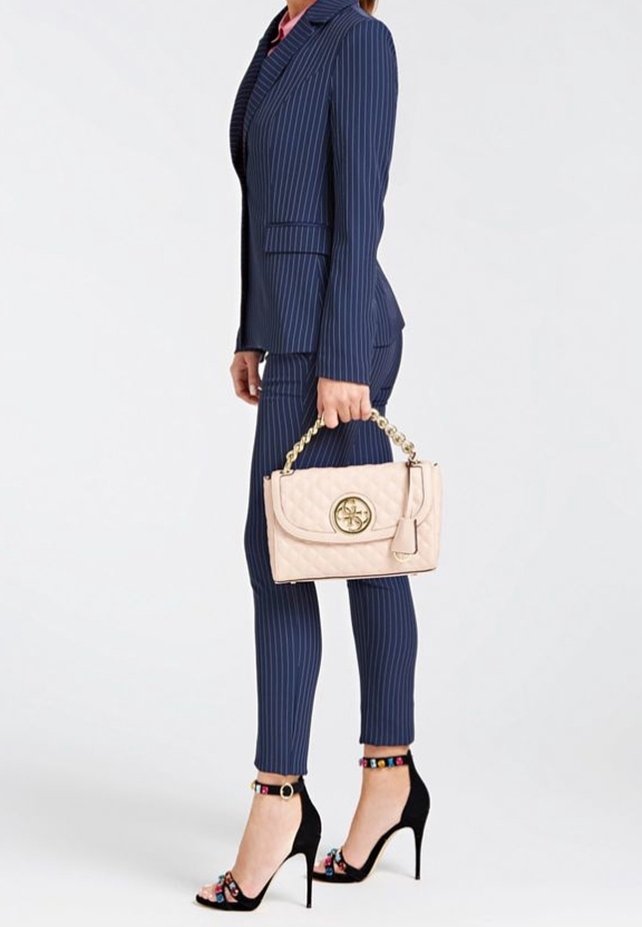Femme en costume bleu marine à fines rayures tenant un sac à main matelassé beige avec une anse chaîne dorée, portant des sandales noires à talons hauts avec des brides colorées à la cheville.