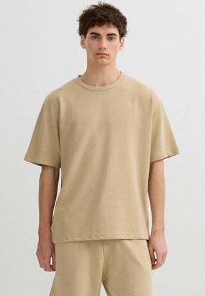 Joven con camiseta de manga corta beige de canalé y pantalones a juego, de pie contra un fondo liso de color gris claro.