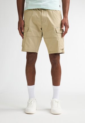 Beige cargoshorts met dubbele zijzakken, zachte katoenen stof en een knielange snit, gecombineerd met witte sokken en sneakers.