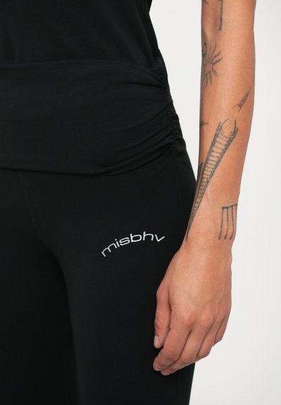 Fekete leggingset viselő személy, amelyen a "misbhv" logó látható a combján, a karján látható tetoválásokkal, és egy fekete ráncolt felsővel.