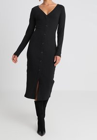 Svart ribbad klänning med långa ärmar, v-ringning och knäppning framtill. Har en slits fram och en figurnära silhuett som når till mitten av vaderna.