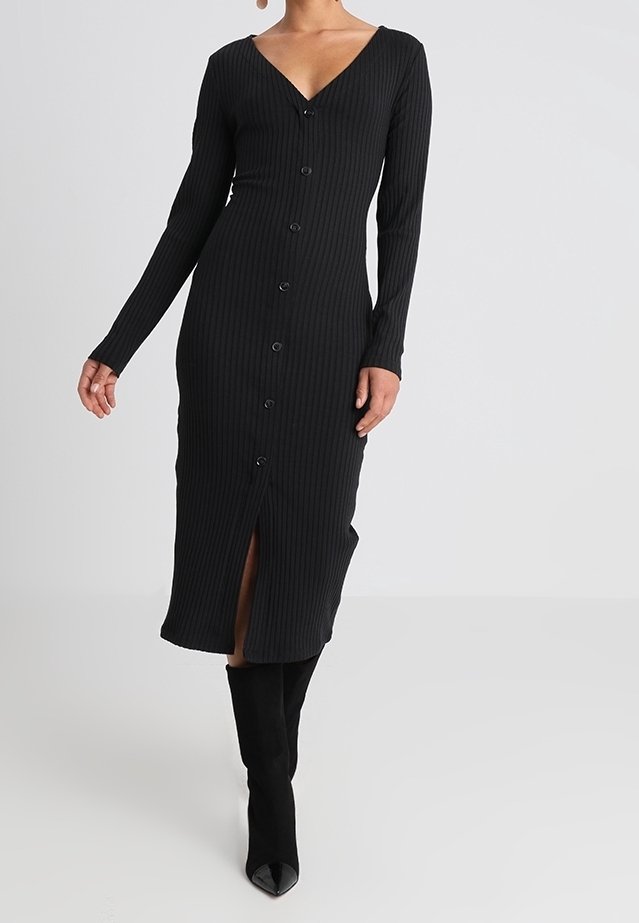 Svart ribbad klänning med långa ärmar, v-ringning och knäppning framtill. Har en slits fram och en figurnära silhuett som når till mitten av vaderna.