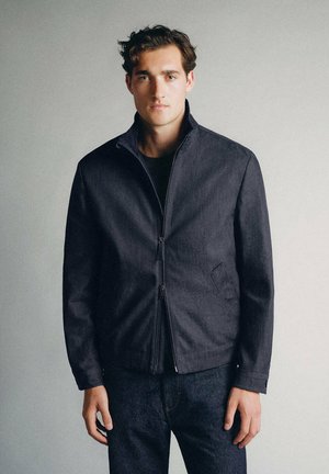 Jeune homme aux cheveux courts et bouclés porte une veste sombre zippée et un pantalon sombre, debout devant un fond gris clair uni.