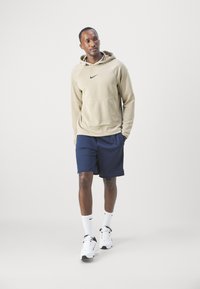 Beež Nike kapuutsiga särk, millel on tekstuuriga disain, tumesinised lühikesed püksid, valged sokid ja must-valged jooksujalatse. Mudel seisab püsti ühetona taustal.