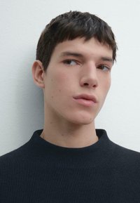 Czarny prążkowany sweter z wysokim kołnierzem o gładkiej fakturze. Tło ma jasny, neutralny kolor.