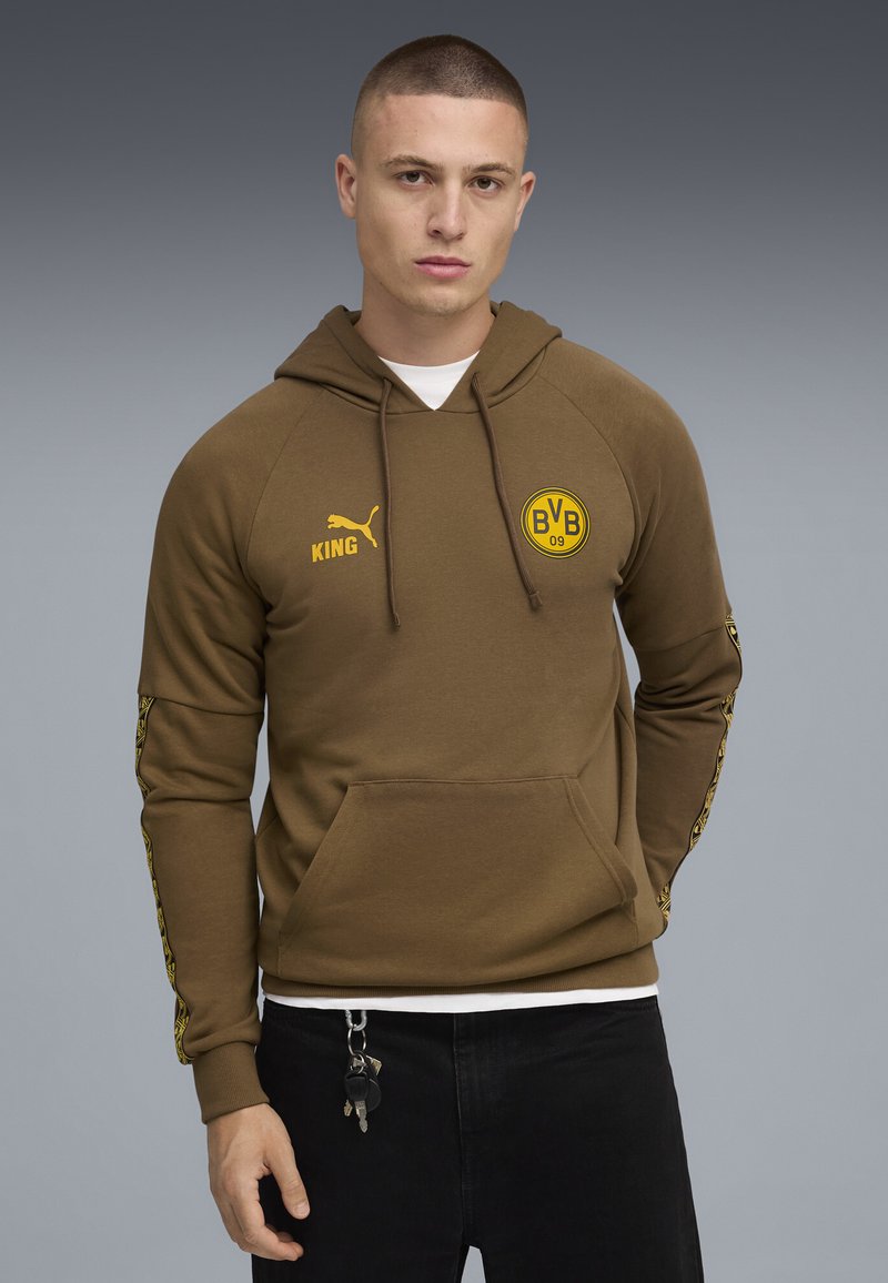 Ung mand iført en brun Puma hoodie med BVB 09 logo, sorte bukser med nøgler hængende, stående foran en grå gradient baggrund.