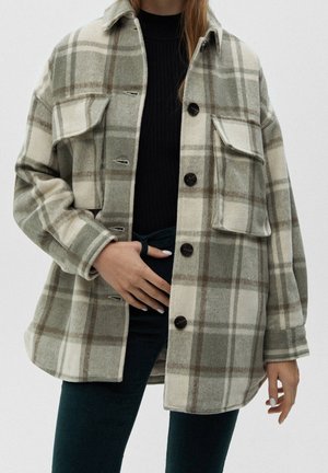 Femme portant une veste à carreaux beige et verte surdimensionnée boutonnée par-dessus un haut noir et un pantalon sombre, une main posée sur la taille.