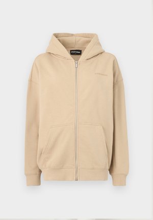 Béžová mikina na zips s kapucňou, prednými vreckami a rebrovými manžetami. Vyrobená z mäkkého materiálu, obsahuje jemné logo na hrudi.