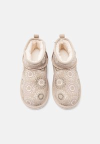 Μπεζ παντόφλες χωρίς κορδόνια με λευκή επένδυση από φλις και κεντητά κυκλικά λουλουδένια σχέδια σε γκρι, λευκό και ανοιχτό μωβ, σε λήψη από ψηλά.