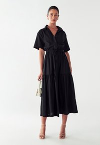 Juoda marškinių tipo suknelė su apykakle, trumpomis rankovėmis ir elastingu liemeniu. Dizaine yra kelių sluoksnių sijonas ir lygus paviršius.