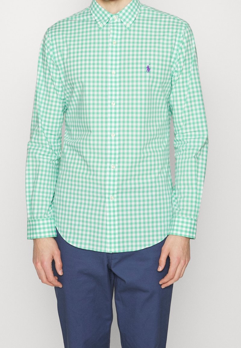 Zelená a bílá košile s kostkovaným vzorem gingham, s límečkem na knoflíky, malým fialovým logem na hrudi a přiléhavými dlouhými rukávy.