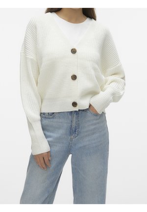 Femme portant un cardigan tricoté blanc épais avec trois grands boutons marron, associé à un jean taille haute bleu clair, main dans la poche.