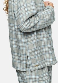 Vêtement en flanelle à carreaux gris avec des accents bleus, présentant une fermeture à boutons, des manches longues et des fentes latérales pour plus de liberté de mouvement.