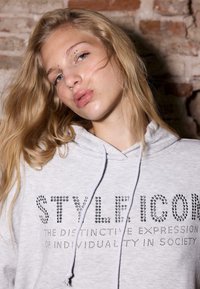 Szara bluza z kapturem z ozdobnym napisem z kryształków, z kapturem na sznurki i miękką fakturą na rustykalnym tle.