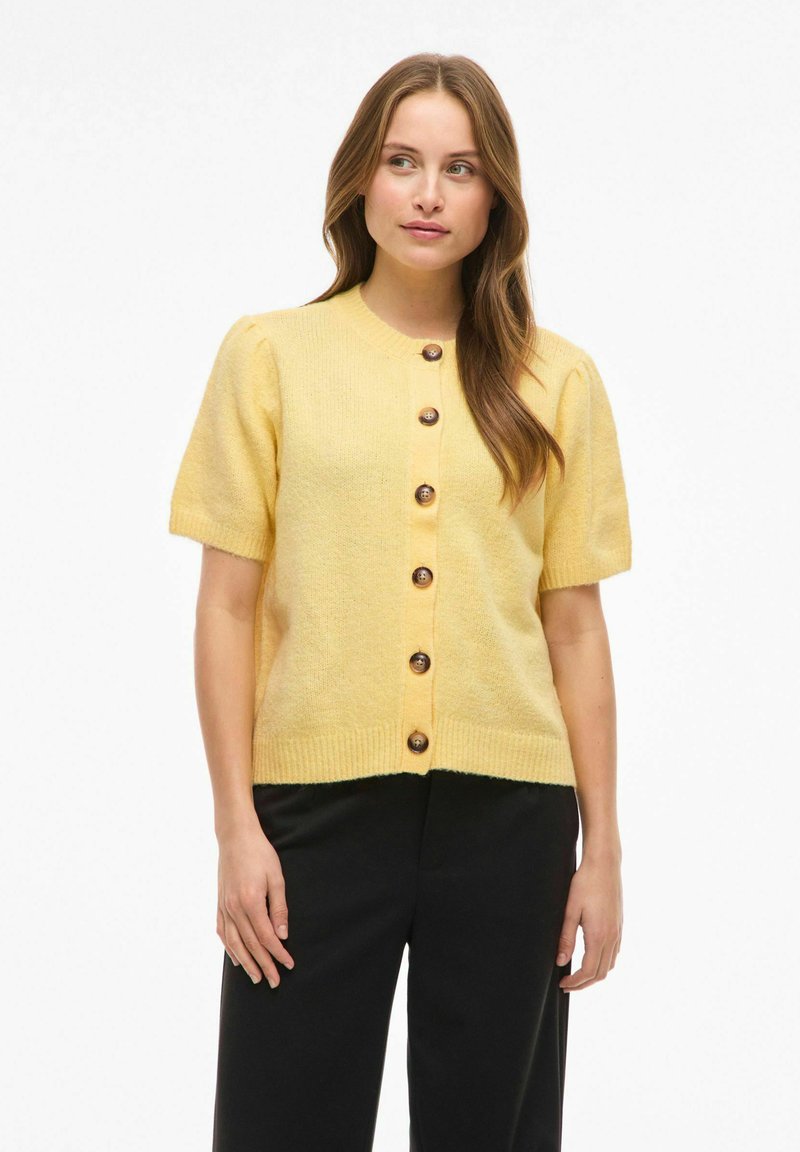 Žlutý krátký cardigan s krátkými rukávy z jemného texturovaného materiálu, s kulatým výstřihem a hnědými knoflíky vpředu. Kombinováno s černými kalhotami.