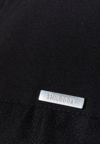 Étiquette rectangulaire métallisée avec le texte "SMILODON" fixée sur un tissu noir texturé en arrière-plan.